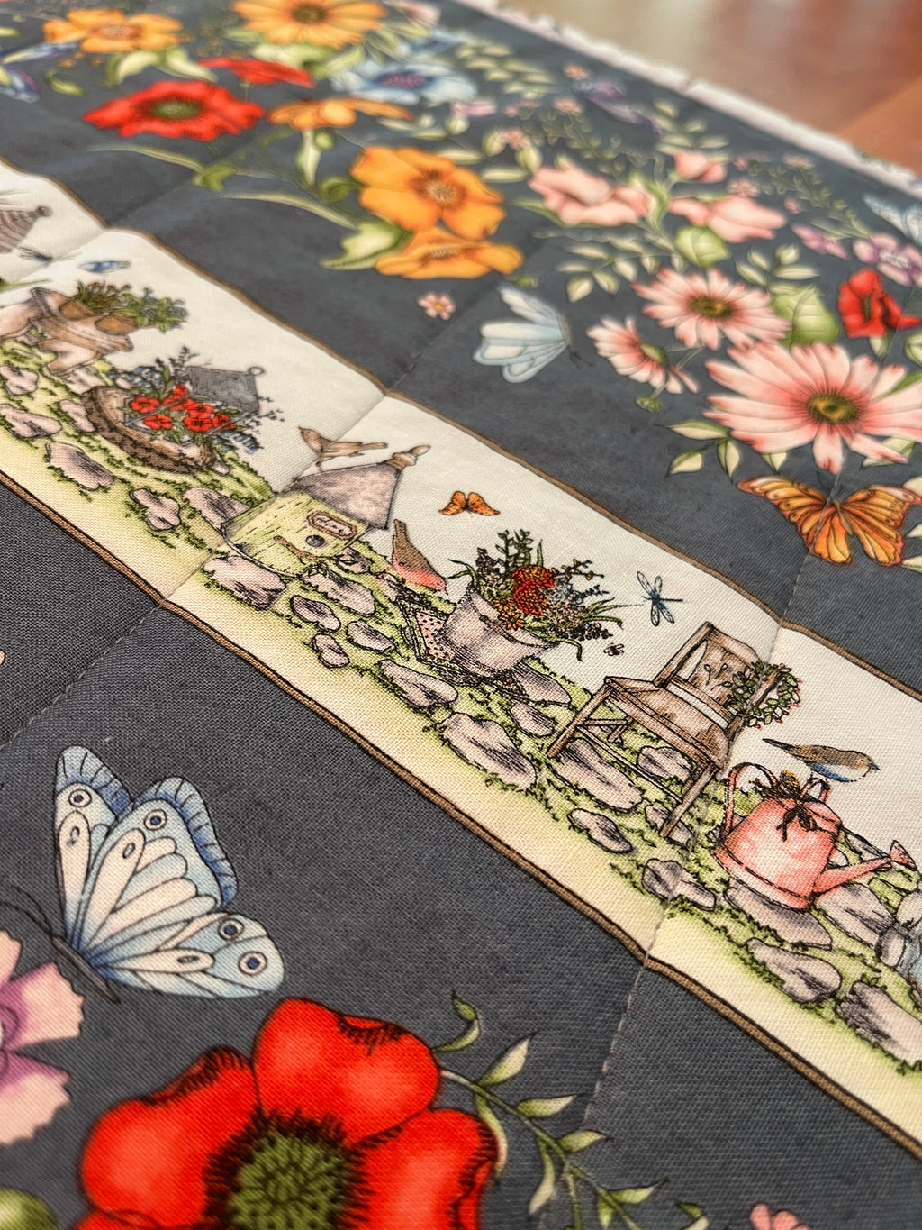 A close-up of a quilted table runner featuring a dark gray background with colorful floral patterns and a central cream-colored border depicting a garden scene with small houses, plants, and a watering can.